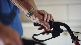 Eine Pflegerin berührt die Hand einer älteren Person, die eine Gehhilfehält (Quelle: IMAGO / Zoonar)