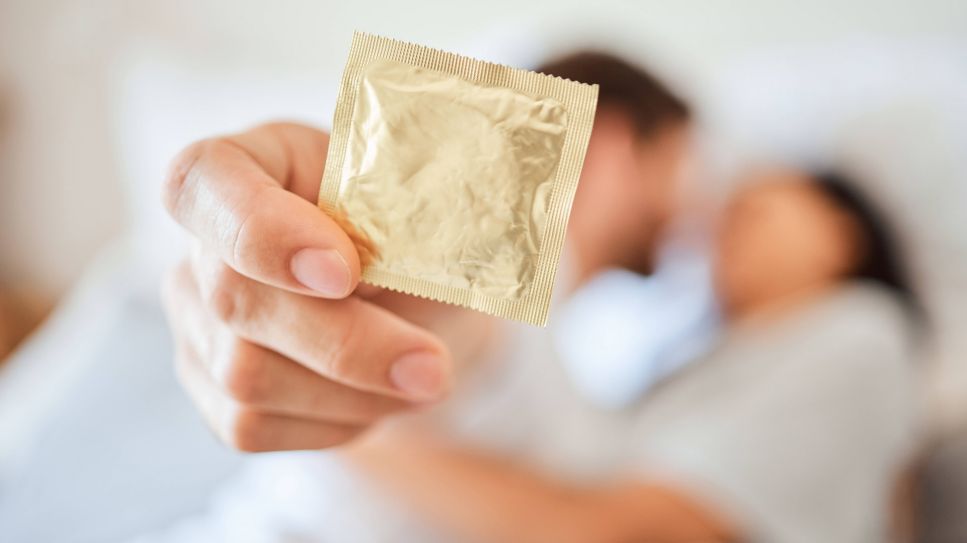 Chlamydien, Symbolbild zeigt Mann, der ein Kondom hält, im Hintergrund unscharf sieht man ihn und eine Frau im Bett (Quelle: imago images / Zoonar)