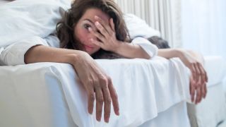 Symbolbild: Eine verkaterte Frau faesst sich in einem Bett liegend in einem Hotelzimmer verschlafen an den Kopf. (Quelle: dpa/Klose)
