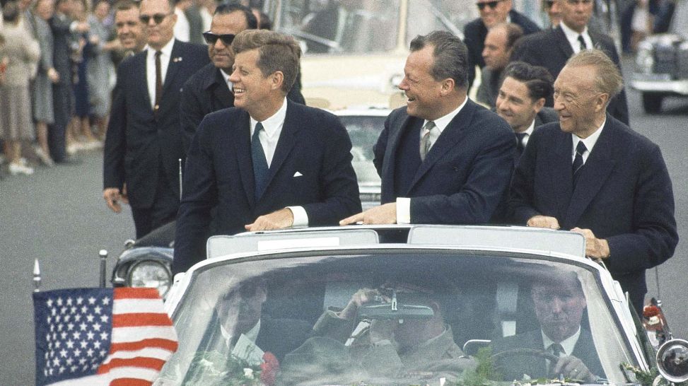 John F. Kennedy, Willy Brandt und Konrad Adenauer stehen im fahrenden Auto (Quelle: picture alliance/AP/Anonymous)