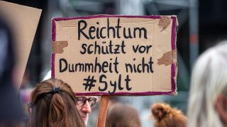 Demonstranten halten am 26.05.2024 ein Schild mit der Aufschrift "Reichtum schützt vor Dummheit nicht #Sylt" hoch (IMAGO / Markus Matzel)