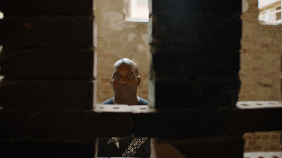 Mann blickt durch einen Spalt in einer Backsteinwand (Quelle: Smiley Baldwin/Flare Film GmbH)