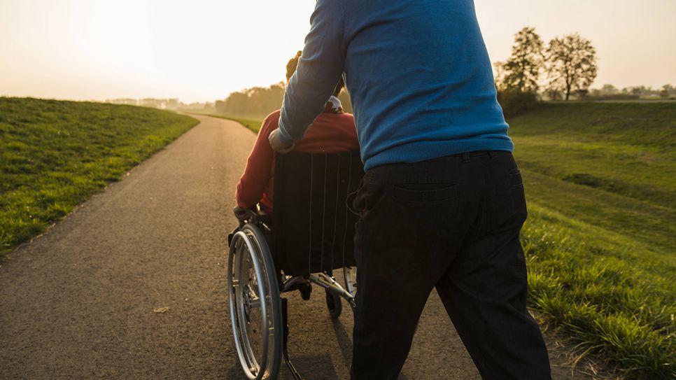 Jemand fährt eine ältere Frau im Rollstuhl auf einem Feldweg entlang (Quelle: imago/Westend61)