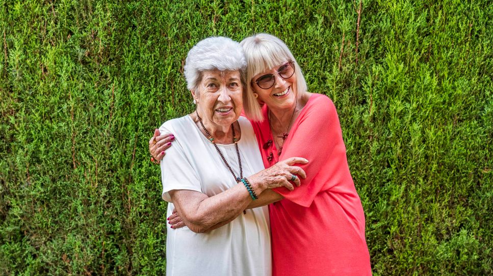 Zwei alte Damen umarmen sich vor einer grünen Hecke (Bild: imago images/Westend61)