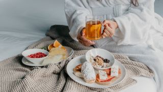Frau mit Tee und Tellern mit Obst und Gebäck auf einem Bett (Bild: imago images/Addictive Stock)