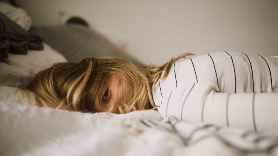 Junge Frau liegt mit offenen Augen bäuchlings auf Bett (Bild: unsplash/Kinga Cichewicz)