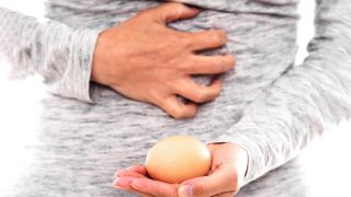 Salmonellen: Frau mit Ei in der Hand fasst sich vor Schmerz an den Bauch (Bild: imago/agefotostock)