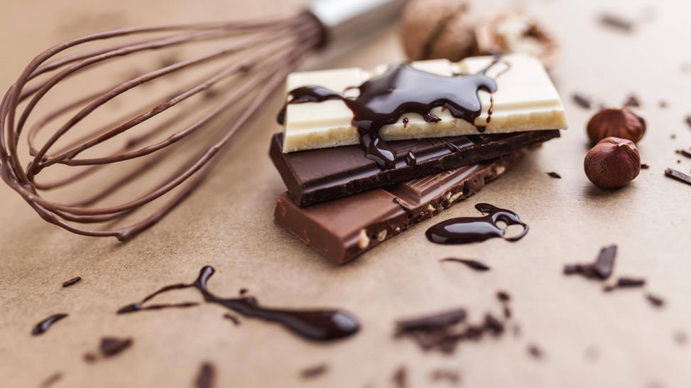 Stückchen verschiedener Schokolade und Rührbesen liegen auf Holztisch (Bild: imago images/Panthermedia)