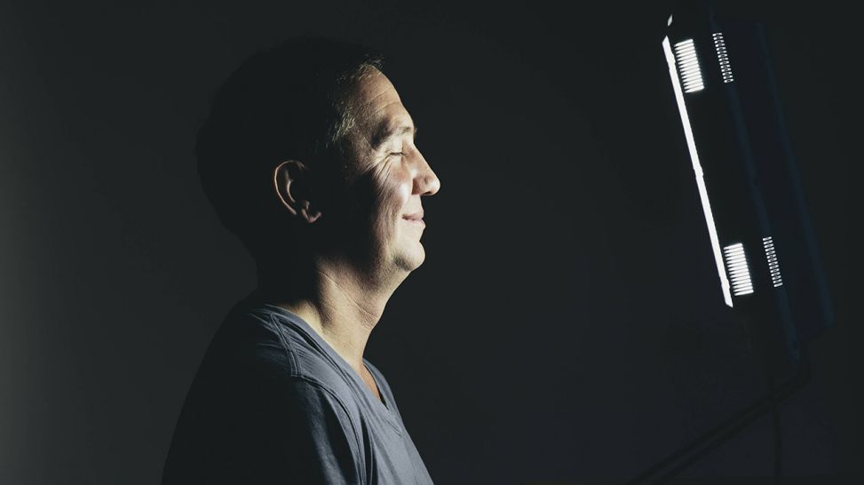 Lächelnder Mann steht vor einer Leuchte zur Lichttherapie (Quelle: imago/Mint Images)