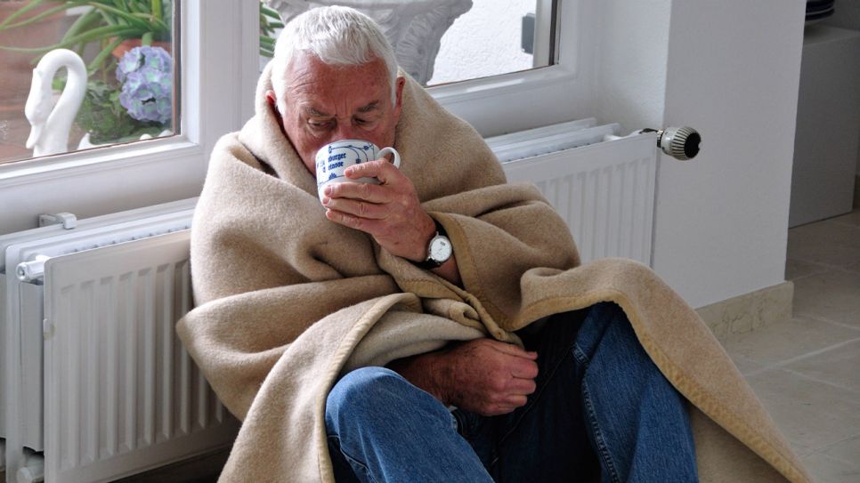 Älterer Mann lehnt mit Decke und Tasse an einem Heizkörper (Quelle: IMAGO / Paul von Stroheim)