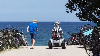 Zwei Männer, davon einer im Rollstuhl, gehen beziehungsweise fahren auf einer Strandpromenade an der Ostsee (Quelle: IMAGO / Jens Koehler)