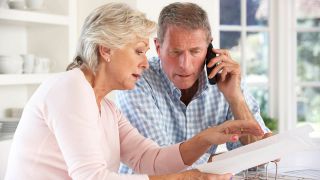 Ein älterer Mann sitzt neben seiner Frau und telefoniert; beide schauen dabei Unterlagen in eniem Ordner an (Quelle: IMAGO / Shotshop)