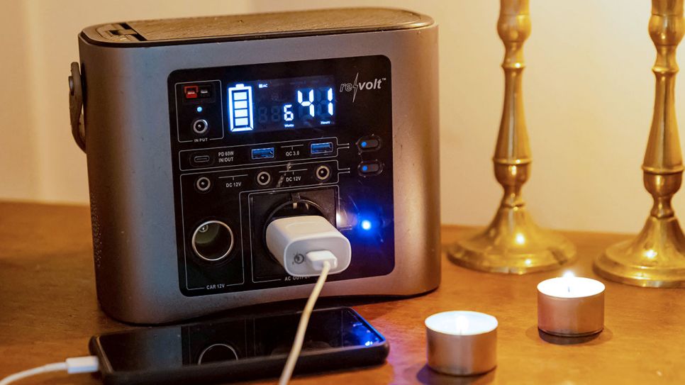 Eine Powerstation steht auf einem Tisch, um sie herum angezündete Teelichter und ein Smartphone, das mit einem Kabel mit der Powerstation verbunden ist (Quelle: IMAGO / Nikito)