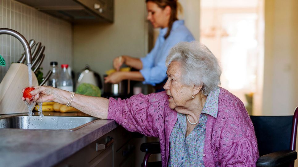Eine im Rollstuhl sitzende Seniorin wäscht eine Tomate am Küchenwaschbecken ab. Im Hintergrund steht eine junge Frau am Herd und kocht (Quelle: IMAGO / Westend61)