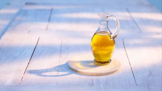 Olivenöl in einer kleinen Karaffe auf einem weißen Tisch, der im Halbschatten im Freien steht (Quelle: IMAGO/Westend61)