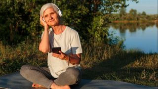 Eine Frau sitzt an einem See und hört sich etwas über Lautsprecher an, das auf dem Smartphone läuft (Quelle: Imago/Westend61)