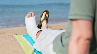 Ein Mann sitzt mit gebrochenem und eingegipsten Fuß auf einem Handtuch am Strand (Quelle: IMAGO / Pond5 Images)