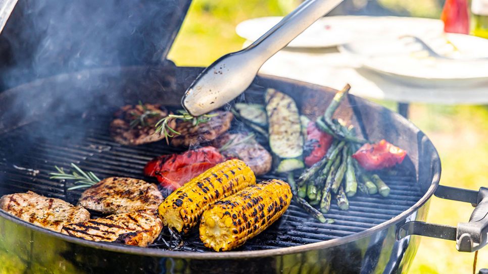 Gemüse und Fleisch wird auf einem rauchendem Kugelgrill mit einer Grillzange auf dem Grillrost sortiert (Quelle: IMAGO / Arnulf Hettrich)