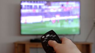 Fernseher mit Fußballspiel und eine Fernbedienung in einer Hand (Quelle: IMAGO / Eibner / Fleig)