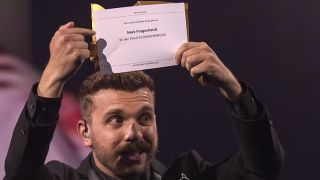 Moderator Edin Hasanovic zeigt auf einen Zettel, der Nora Fingscheidt zur Gewinnerin des Preises für die beste Regie im Film "Systemsprenger" macht (Bild: rbb)