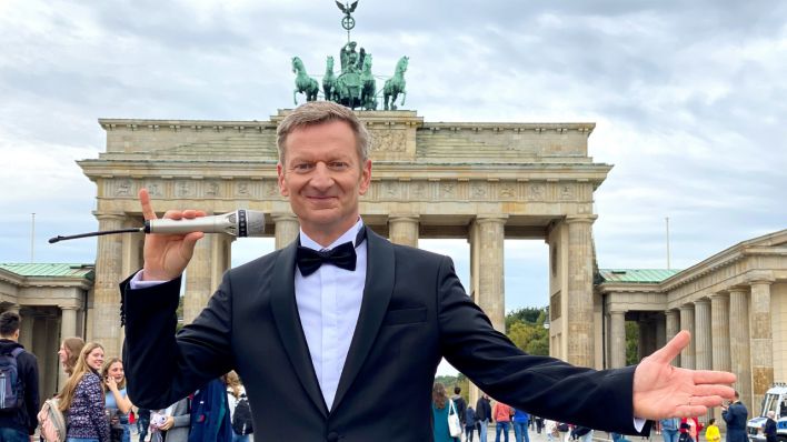 Michael Kessler trägt einen schwarzen Anzug und steht mit Mikro vor dem Brandenburger Tor
